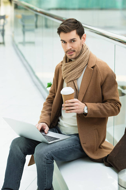 knappe man met laptop zittend op de Bank, de holding van wegwerp cup en de camera kijken - Foto, afbeelding