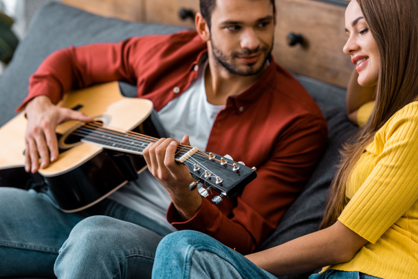 cropped view of man sitting on sofa and playing guitar for smiling girlfriend - Fotografie, Obrázek