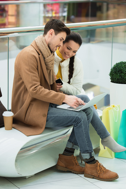 smiling girl sitting on bench with handsome man pointing with finger on laptop screen in shopping mall - Photo, Image