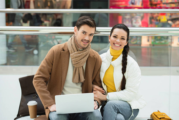 handsome man with laptop and beautiful girl sitting on bench and looking at camera - Foto, Imagem