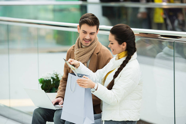 homme souriant avec ordinateur portable et fille avec sac à provisions assis sur le banc et regardant les achats dans le centre commercial
 - Photo, image