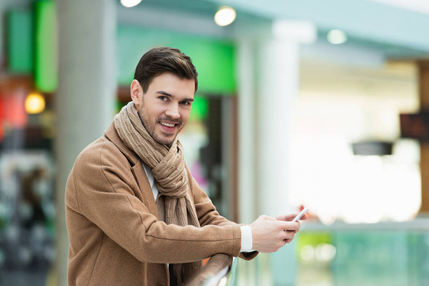 hombre sonriente con ropa de abrigo sosteniendo el teléfono inteligente y mirando a la cámara
 - Foto, Imagen