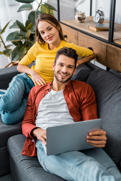 young attractive couple sitting on sofa with laptop and smiling while looking at camera - 写真・画像