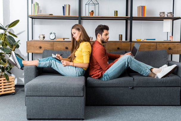 young smiling woman with notebook sitting back to back on sofa with man using laptop - Foto, Imagem