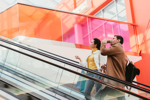 handsome man drinking coffee from disposable cup and attractive girl with shopping bags on escalator in mall - Photo, Image