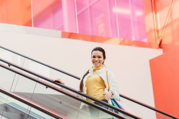 attractive girl with shopping bags smiling and looking at camera on escalator - Foto, afbeelding