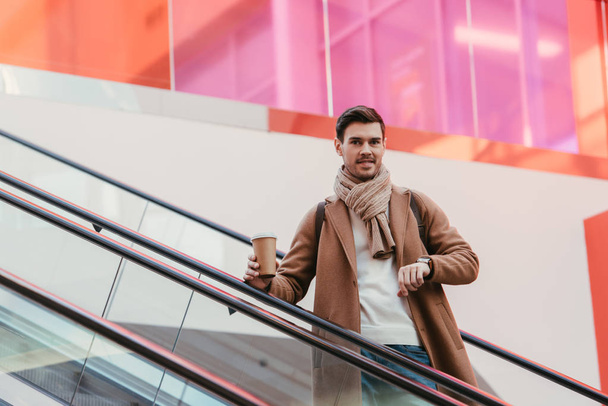 handsome man in warm clothing holding disposable cup, smiling and looking at camera on escalator - Фото, зображення