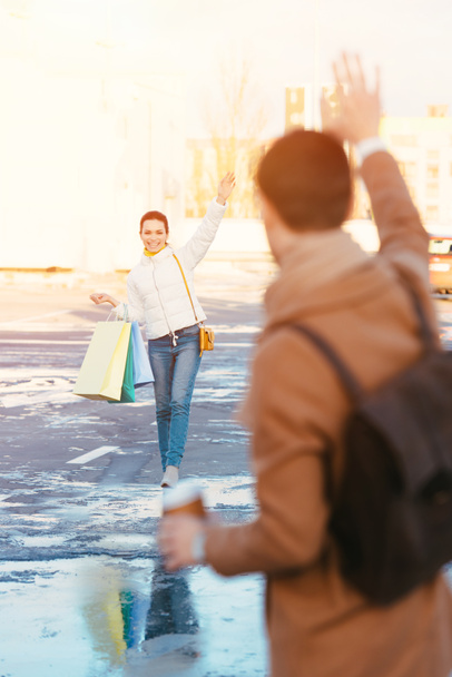 chica atractiva con bolsas de compras saludando con la mano al novio en el aparcamiento
 - Foto, Imagen