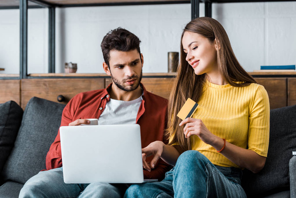 attractive woman holding credit card while boyfriend sitting with laptop - Φωτογραφία, εικόνα