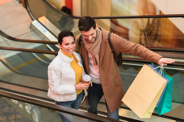 hombre guapo con bolsas de compras y chica atractiva con taza desechable subiendo en escaleras mecánicas
 - Foto, imagen