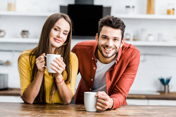 selective focus of cheerful couple holding cups with drinks in kitchen - Foto, Imagen
