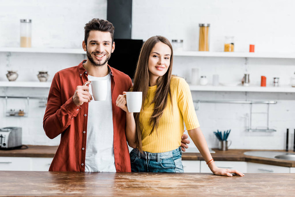 cheerful couple standing in kitchen and smiling with cups  - Фото, изображение