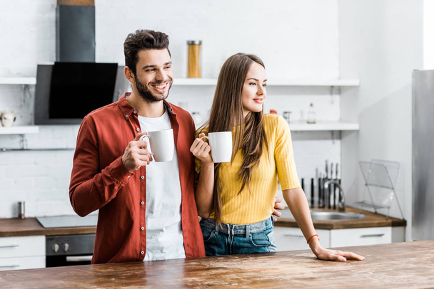 cheerful couple standing in kitchen with cups  - Valokuva, kuva