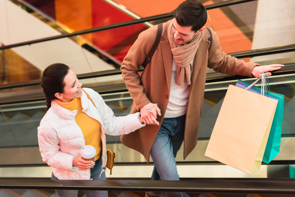 hombre sonriente con bolsas de compras y hermosa chica con taza desechable mirándose el uno al otro en escaleras mecánicas
 - Foto, Imagen