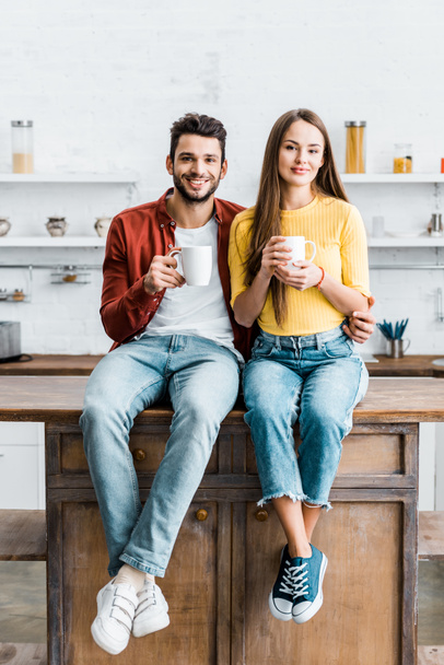 cheerful couple sitting on wooden table in kitchen and holding cups - Foto, Bild