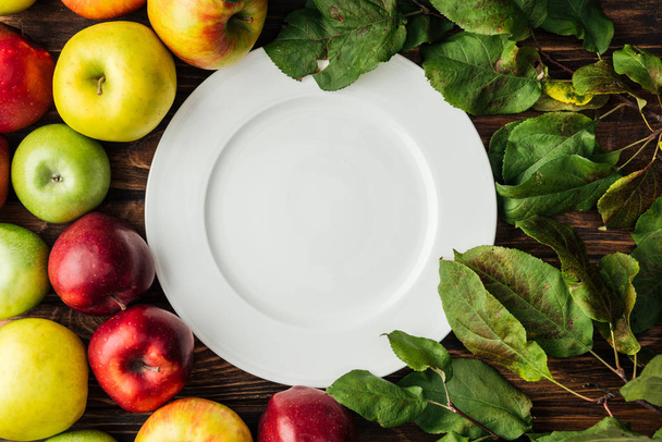 vista superior de la placa blanca, manzanas maduras multicolores y ramas con hojas en mesa de madera
 - Foto, Imagen