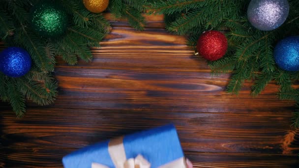 Female hands puts gift wrapped in blue paper with bow on top on wooden table and then takes it. Christmas decorations - flashing garland lights, fir branches, bauble. Flat lay. - Footage, Video