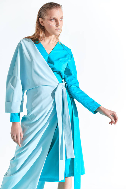 Fashion portrait of young blonde woman with blue eyes and painted white lines on face, dressed in turquoise dress. White background - Foto, imagen