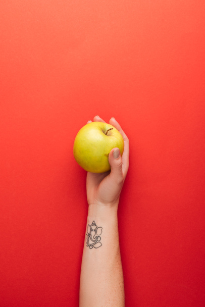 vista superior de la mano femenina tatuada sosteniendo gran manzana deliciosa dorada sobre fondo rojo
 - Foto, Imagen
