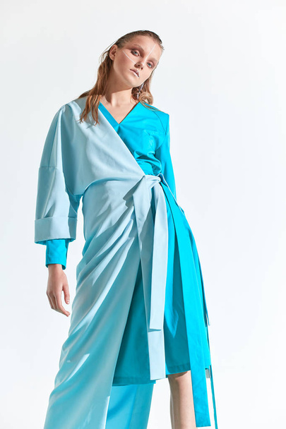 Fashion portrait of young blonde woman with blue eyes and painted white lines on face, dressed in turquoise dress. White background - Photo, Image