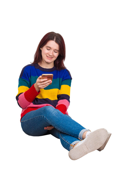 Full length portrait of carefree attractive young woman, smiling broadly sitting on the floor browse phone. Casual girl wearing jeans and sweater isolated over white. - Foto, Bild