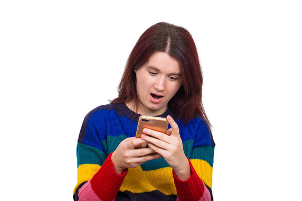 junges Weibchen mit angenehm geöffnetem Mund blickt mit erschrockenem Gesichtsausdruck auf Handy, liest überraschende Nachrichten auf Webseite, isoliert über weißem Hintergrund. - Foto, Bild