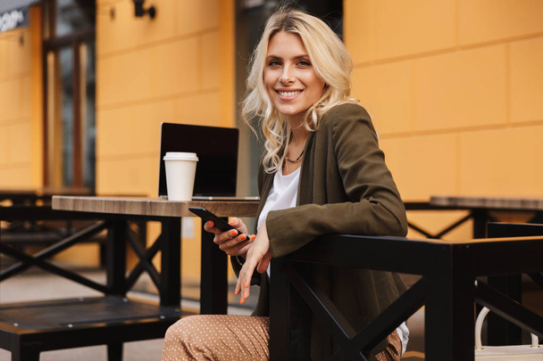 Portrait of beautiful woman 20s holding cell phone while sitting in cafe outdoor with takeaway coffee and silver laptop - Фото, изображение