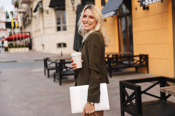 Image of blonde woman wearing jacket holding takeaway coffee and laptop while walking through city street - Foto, afbeelding