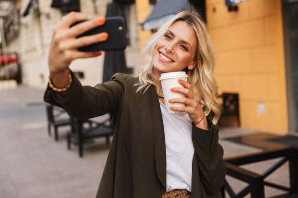 Image of beautiful woman holding takeaway coffee in paper cup and taking selfie while walking through city street - Фото, изображение