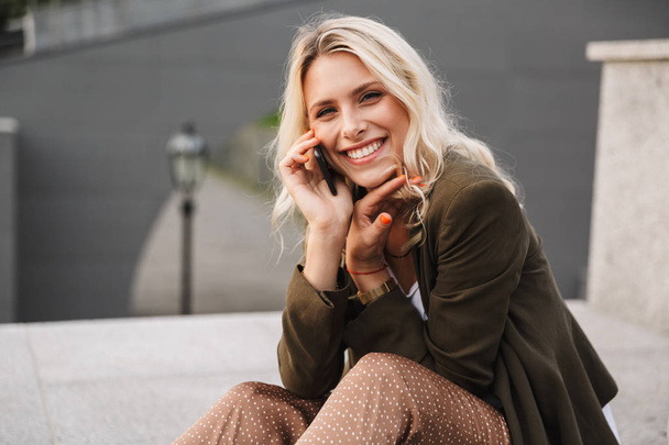 笑顔の女性の画像 20 代のスマート フォンでは話しながら屋外コンクリートの欄干の上に座って - 写真・画像