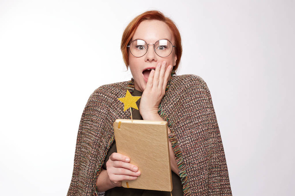 Attractive young emotional Caucasian freckled female looks with great surprise at camera, keeps mouth widely opened, not expected to receive such news, wearing  jacket on shoulders, holding book. - Photo, Image