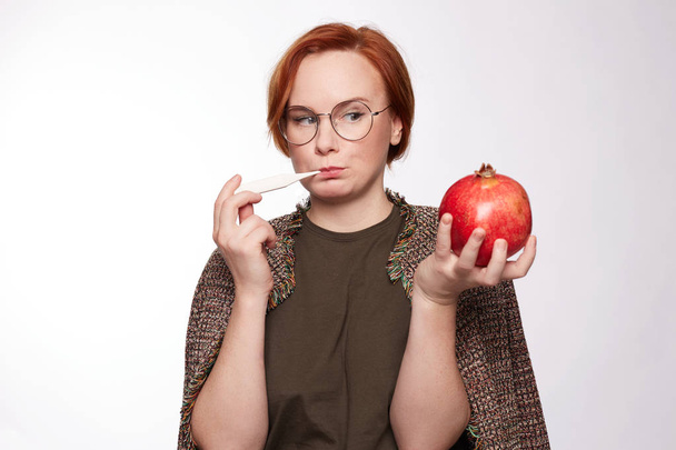 Young freckled redhead woman uses thermometer, feels unwell, has flu. Sick female has fever or allergic, tired look, needs good rest, holds pomegranate to get vitamins. Unhealthy lifestyle, infection. - 写真・画像