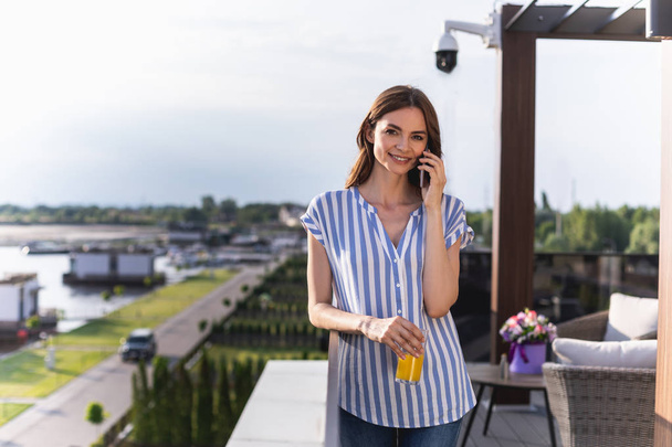 Waist up portrait of young woman standing on balcony and speaking on handy. She looking at camera with smile. Copy space on left side - 写真・画像