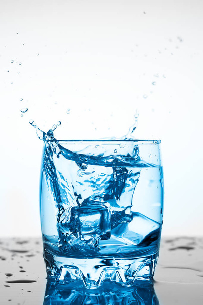 water splash in a glass with a piece of ice on a white background, a piece of ice falling into glass Cup of water, splashes of water flying in all directions - Photo, Image