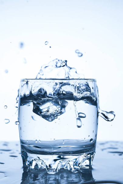 water splash in a glass with a piece of ice on a white background, a piece of ice falling into glass Cup of water, splashes of water flying in all directions - Foto, Imagem