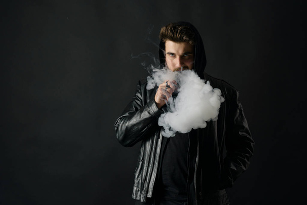 Man with vaping mod exhaling steam at black studio background. Bearded guy smoking e-cigarette to quit tobacco. Vapor and alternative nicotine free smoking concept - Фото, изображение
