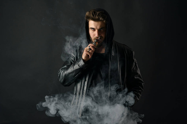 Man with vaping mod exhaling steam at black studio background. Bearded guy smoking e-cigarette to quit tobacco. Vapor and alternative nicotine free smoking concept - 写真・画像