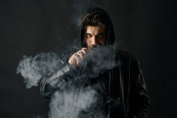 Man with vaping mod exhaling steam at black studio background. Bearded guy smoking e-cigarette to quit tobacco. Vapor and alternative nicotine free smoking concept - Fotografie, Obrázek