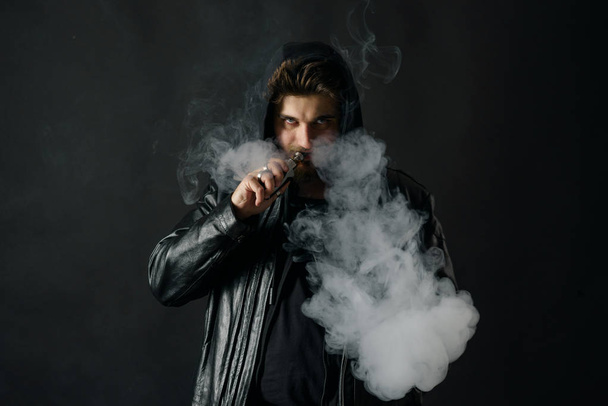 Man with vaping mod exhaling steam at black studio background. Bearded guy smoking e-cigarette to quit tobacco. Vapor and alternative nicotine free smoking concept - Photo, Image