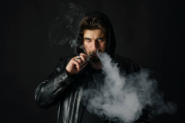 Man with vaping mod exhaling steam at black studio background. Bearded guy smoking e-cigarette to quit tobacco. Vapor and alternative nicotine free smoking concept - Fotó, kép