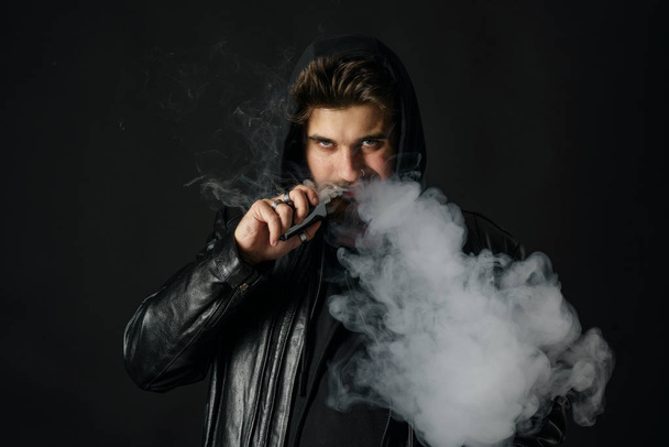 Man with vaping mod exhaling steam at black studio background. Bearded guy smoking e-cigarette to quit tobacco. Vapor and alternative nicotine free smoking concept - Foto, imagen