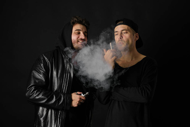 Bad habit, smoking in a public place. Two Young man blowing smoke to join it in one cloud at black studio background. Friends and vape addiction concept.  - Foto, Imagen