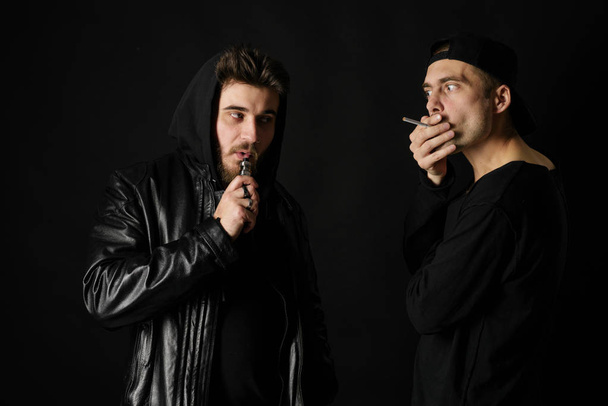 Bad habit, smoking in a public place. Two Young man blowing smoke to join it in one cloud at black studio background. Friends and vape addiction concept.  - Fotografie, Obrázek