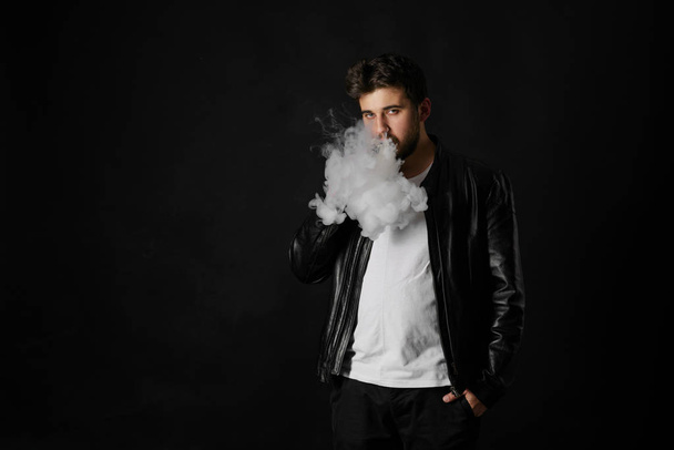Man with vaping mod exhaling steam at black studio background. Bearded guy smoking e-cigarette to quit tobacco. Vapor and alternative nicotine free smoking concept - 写真・画像
