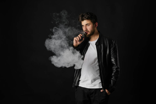 Man with vaping mod exhaling steam at black studio background. Bearded guy smoking e-cigarette to quit tobacco. Vapor and alternative nicotine free smoking concept - Zdjęcie, obraz