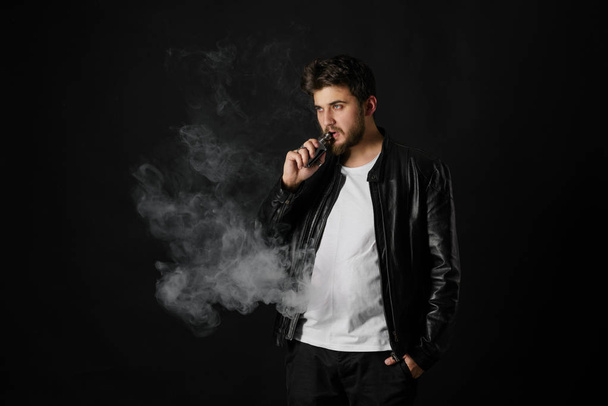 Man with vaping mod exhaling steam at black studio background. Bearded guy smoking e-cigarette to quit tobacco. Vapor and alternative nicotine free smoking concept - Zdjęcie, obraz
