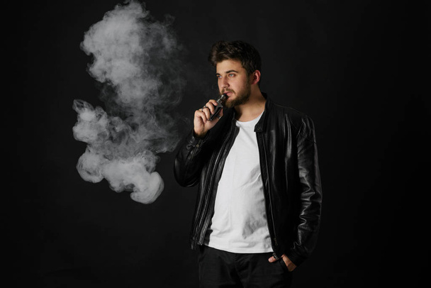 Man with vaping mod exhaling steam at black studio background. Bearded guy smoking e-cigarette to quit tobacco. Vapor and alternative nicotine free smoking concept - Foto, immagini