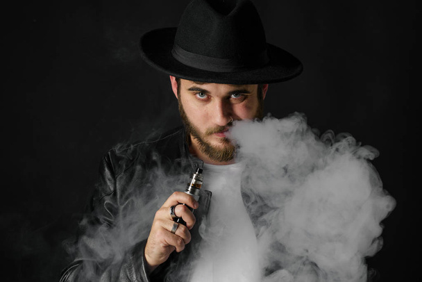 Man with vaping mod exhaling steam at black studio background. Bearded guy smoking e-cigarette to quit tobacco. Vapor and alternative nicotine free smoking concept - Valokuva, kuva