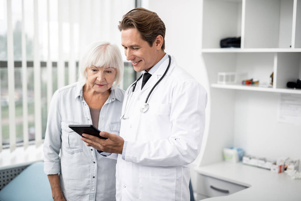 Curious aged woman looking at the screen of tablet and doctor smiling - Foto, Imagem