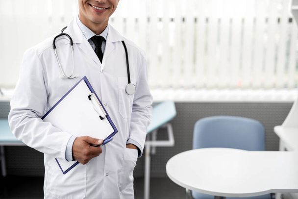 Médico emotivo alegre vestindo roupas médicas brancas e sorrindo enquanto segurando prancheta e tendo estetoscópio em seu pescoço
 - Foto, Imagem